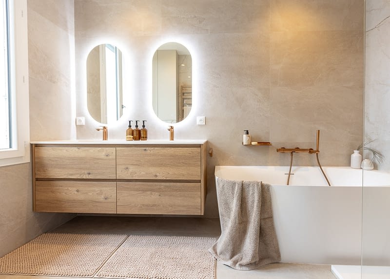 Vue d’ensemble de la salle de bain beige, avec meuble suspendu en bois, double vasque, deux miroirs lumineux et baignoire blanche.